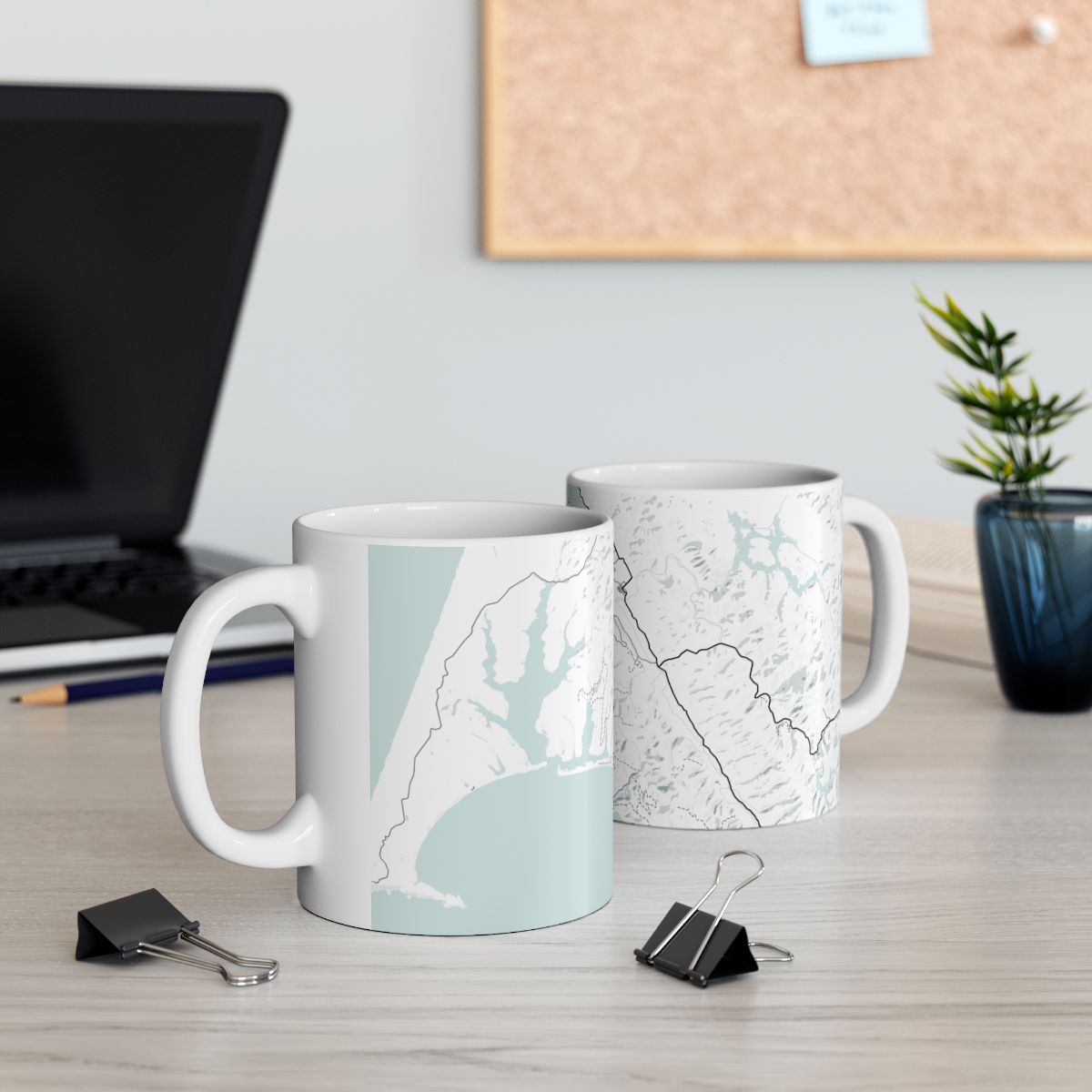 Two Atlas & Ink mugs with map designs on a desk with office supplies and a plant.